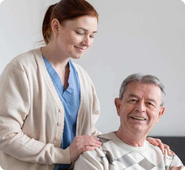 A nurse in a blue uniform and beige cardigan smiles while gently placing a hand on the shoulder of an elderly man in a light-colored, diamond-patterned sweater. They are in a simple room with a couch in the background.