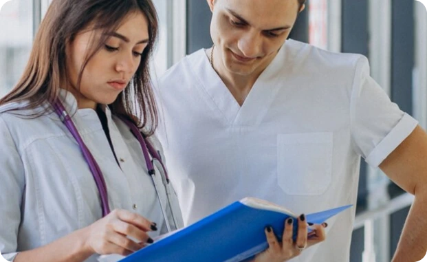 Two healthcare professionals in white scrubs, one with a stethoscope around their neck, are reviewing a blue patient chart together in a clinical setting.