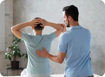 A healthcare professional in a blue shirt examines a patient's shoulder in a clinical setting, with the patient sitting on a table and a potted plant in the background.