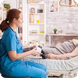 A nurse in blue scrubs takes the blood pressure of a patient lying in a hospital bed, in a cozy room with shelves, books, plants, and framed pictures on the wall.