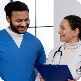 A nurse in blue scrubs smiles while discussing with a doctor in a white coat holding a clipboard and wearing a stethoscope, in a clinical setting with large windows in the background.