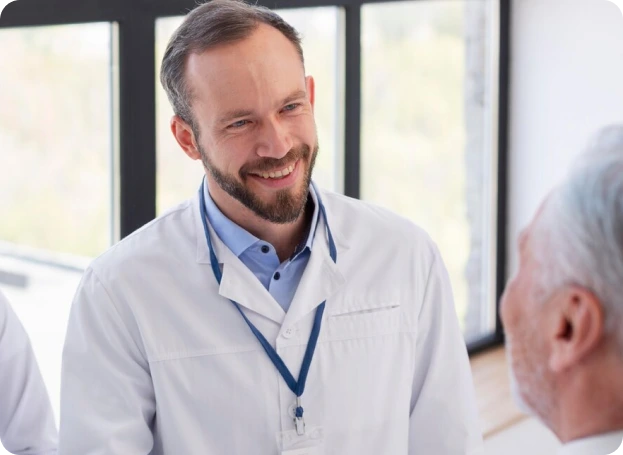 A doctor in a white lab coat with a blue lanyard smiles while engaging in conversation with an elderly person in a room with large windows.