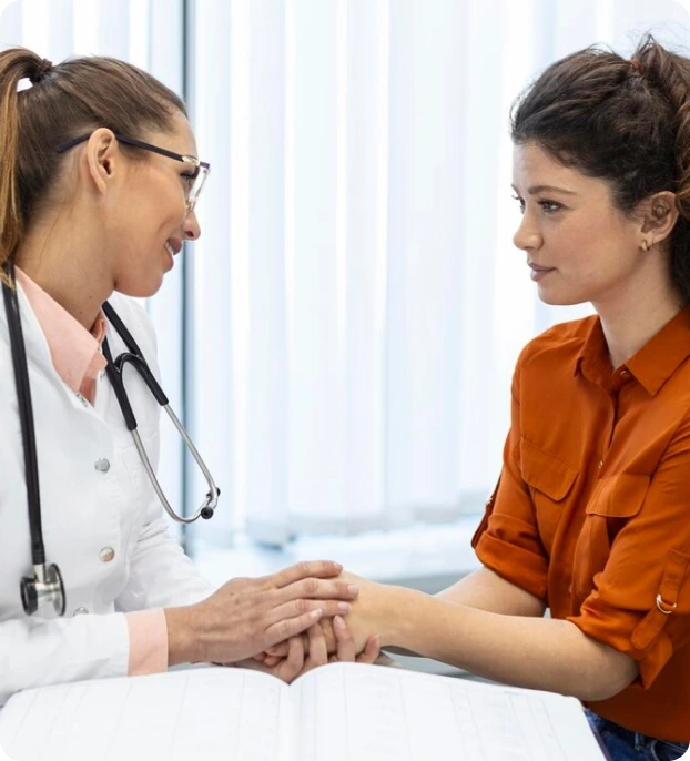 A doctor in a white coat with a stethoscope around their neck holds hands with a patient in an orange shirt, sitting across from each other in a clinical setting with vertical blinds in the background. An open chart lies on the table between them.