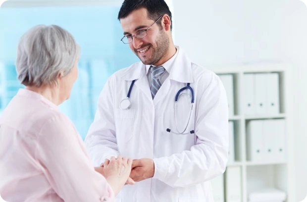 A doctor in a white coat with a stethoscope around their neck holds hands with an elderly woman in a light pink top, both smiling in a clinical setting with shelves and files in the background.