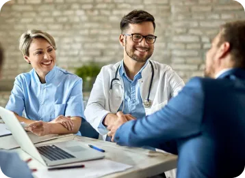 A healthcare professional in a white coat with a stethoscope shakes hands with a person in a suit across a table, while another professional in blue scrubs smiles in the background. A laptop and papers are on the table in a modern office setting with a brick wall.