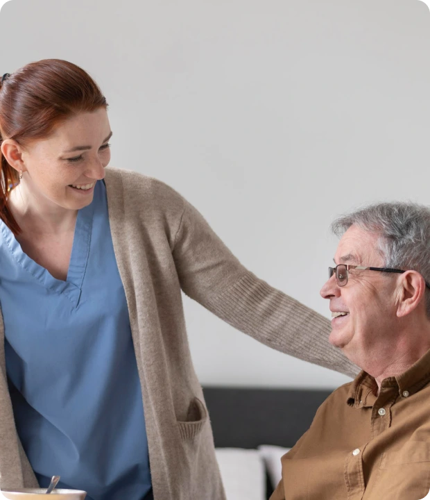 A nurse in a blue uniform and beige cardigan smiles while gently touching the shoulder of an elderly man in a brown shirt, who is sitting on a bed. The setting appears to be a simple room with a bed and a bowl with a spoon on a nearby surface.