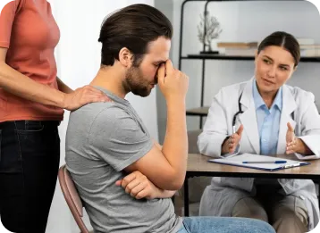 A healthcare professional in a white coat consults with a distressed patient sitting at a table, while another person in a red shirt offers support by placing a hand on the patient's shoulder. A clipboard and papers are on the table in a clinical setting with shelves and decor in the background.