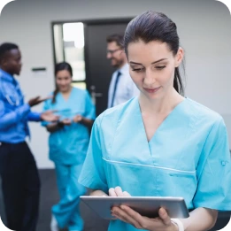 A nurse in blue scrubs uses a tablet in a clinical setting, while other medical professionals in blue scrubs and a doctor in a white coat discuss in the background near a door.