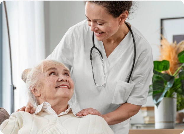 A nurse in a white uniform with a stethoscope around her neck smiles warmly while gently supporting an elderly woman sitting on a couch. The elderly woman, dressed in a light-colored sweater, looks up at the nurse with a smile. They are in a cozy room with plants and framed pictures in the background.