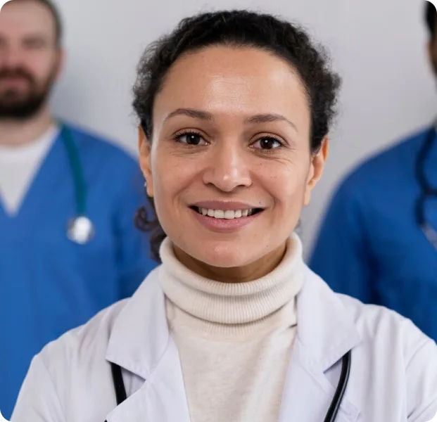 A smiling healthcare professional in a white coat with a stethoscope around their neck stands in the foreground, with two other professionals in blue scrubs blurred in the background.
