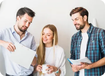 Three professionals in casual attire collaborate in an office setting. One holds a clipboard, another holds a mug and a notebook, and the third holds a book, all smiling and engaged in discussion against a light background.