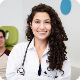 A doctor in a white coat with a stethoscope around their neck stands with arms crossed, smiling confidently in a clinical setting. Another person in a brown shirt is seated in the background near a green chair.