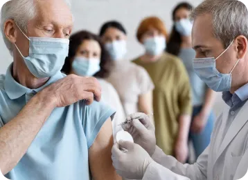 A healthcare professional in a white coat and gloves administers a vaccine to an elderly person in a blue shirt, while several others wearing face masks observe in the background.