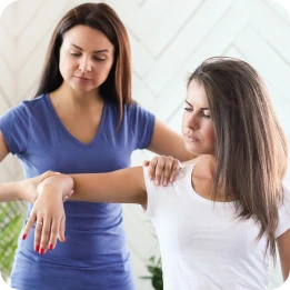 A physical therapist in a blue uniform assists a patient in a white shirt with a shoulder exercise, holding the patient's arm in a clinical room with plants in the background.