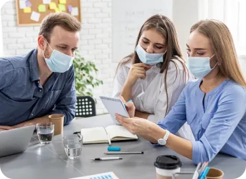 Three professionals wearing face masks collaborate at a table in an office with a brick wall and bulletin board in the background. They review a notebook, with a laptop, coffee cups, and papers on the table.