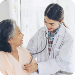 A doctor in a white coat uses a stethoscope to check the heartbeat of an elderly woman in a light pink top, both smiling in a clinical setting with vertical blinds in the background.