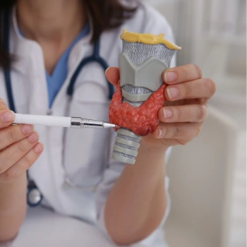 A person wearing a white lab coat holds a model of a thyroid gland, using a pen to point at it, symbolizing a thyroid test or examination. The thyroid model is red with a gray and yellow section above it, likely representing the larynx and trachea. The background is blurred, suggesting a clinical setting.