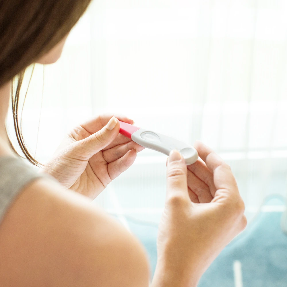 The image shows a person with long brown hair holding a pregnancy test stick with a red and white handle, viewed from behind.