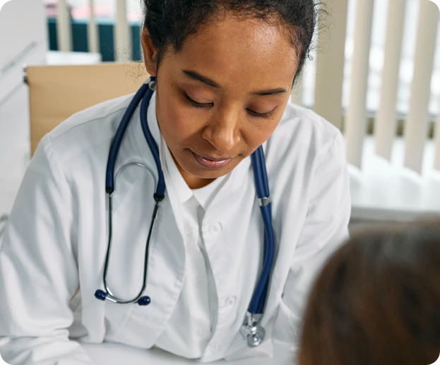 A person in a white lab coat with a stethoscope around their neck is focused on a patient, sitting at a desk in a room with beige blinds and a chair in the background.