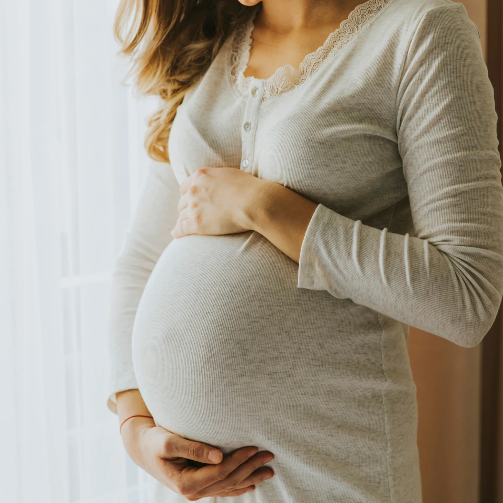 The image shows a pregnant woman with long wavy hair, gently holding her baby bump. The background features a softly lit room with sheer curtains and a warm-toned wall.
