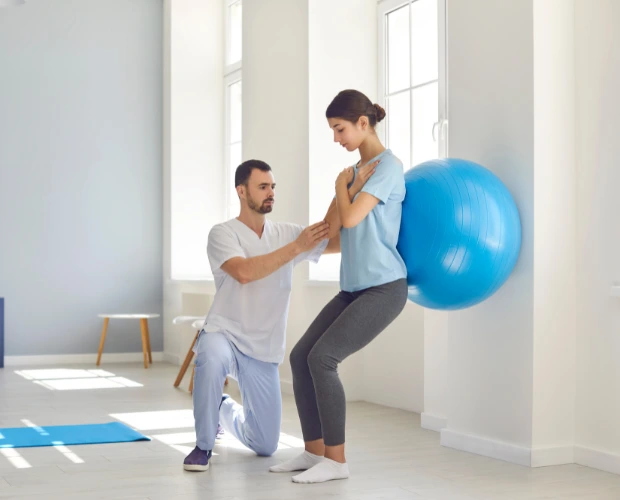 A person in a white medical uniform kneels beside another person, assisting with a physical therapy exercise. The person in the blue shirt is squatting while leaning against a large blue exercise ball on the wall.