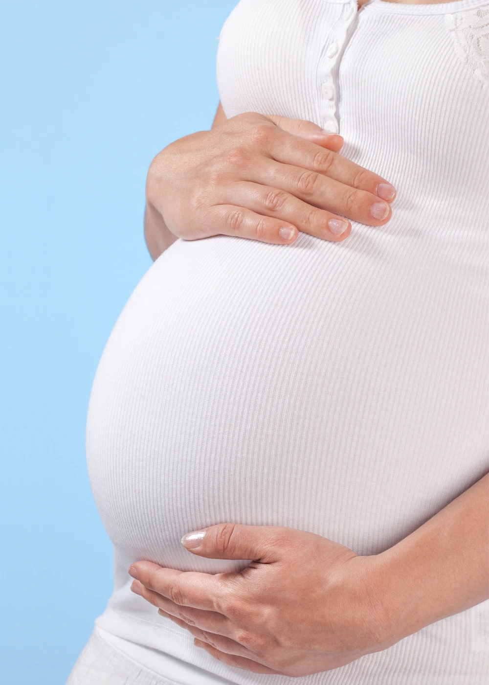 Pregnant woman in a white ribbed shirt holding her baby bump against a light blue background.