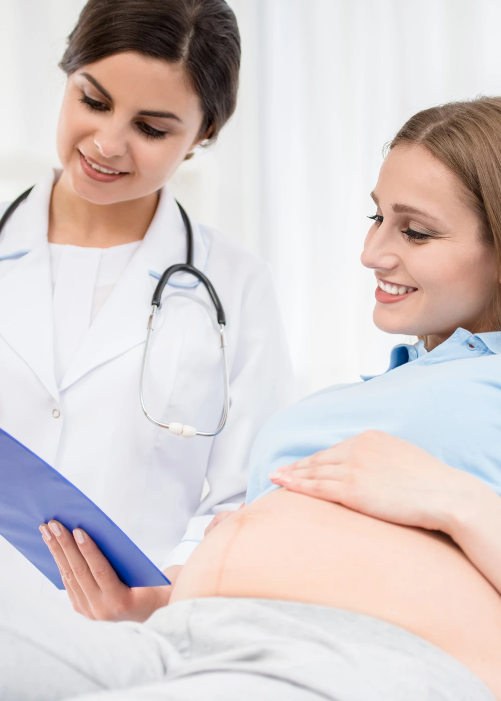 A doctor in a white coat with a stethoscope, holding a blue medical chart, consulting with a pregnant woman who is lying down and resting her hand on her baby bump.