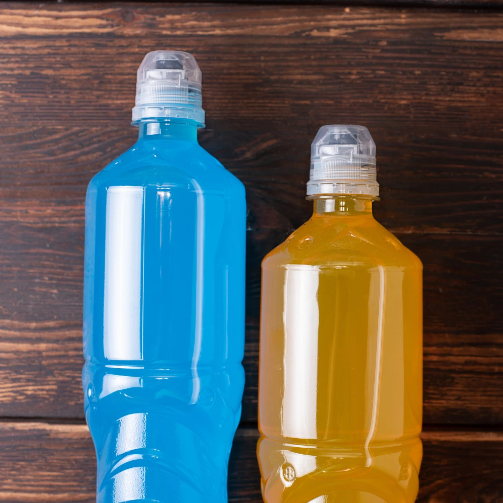 Two plastic bottles of electrolyte drinks, one blue and one orange, with white caps, placed on a dark wooden surface.