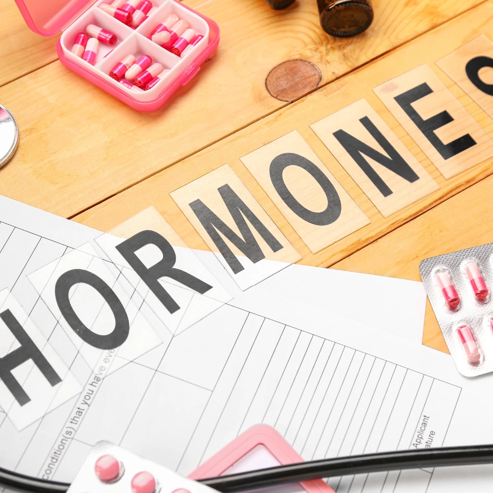 A wooden table with a pink pill organizer containing various pills, a blister pack of pink and white capsules. On the table, large letters spelling "HORMONE" are arranged, placed over a medical form.