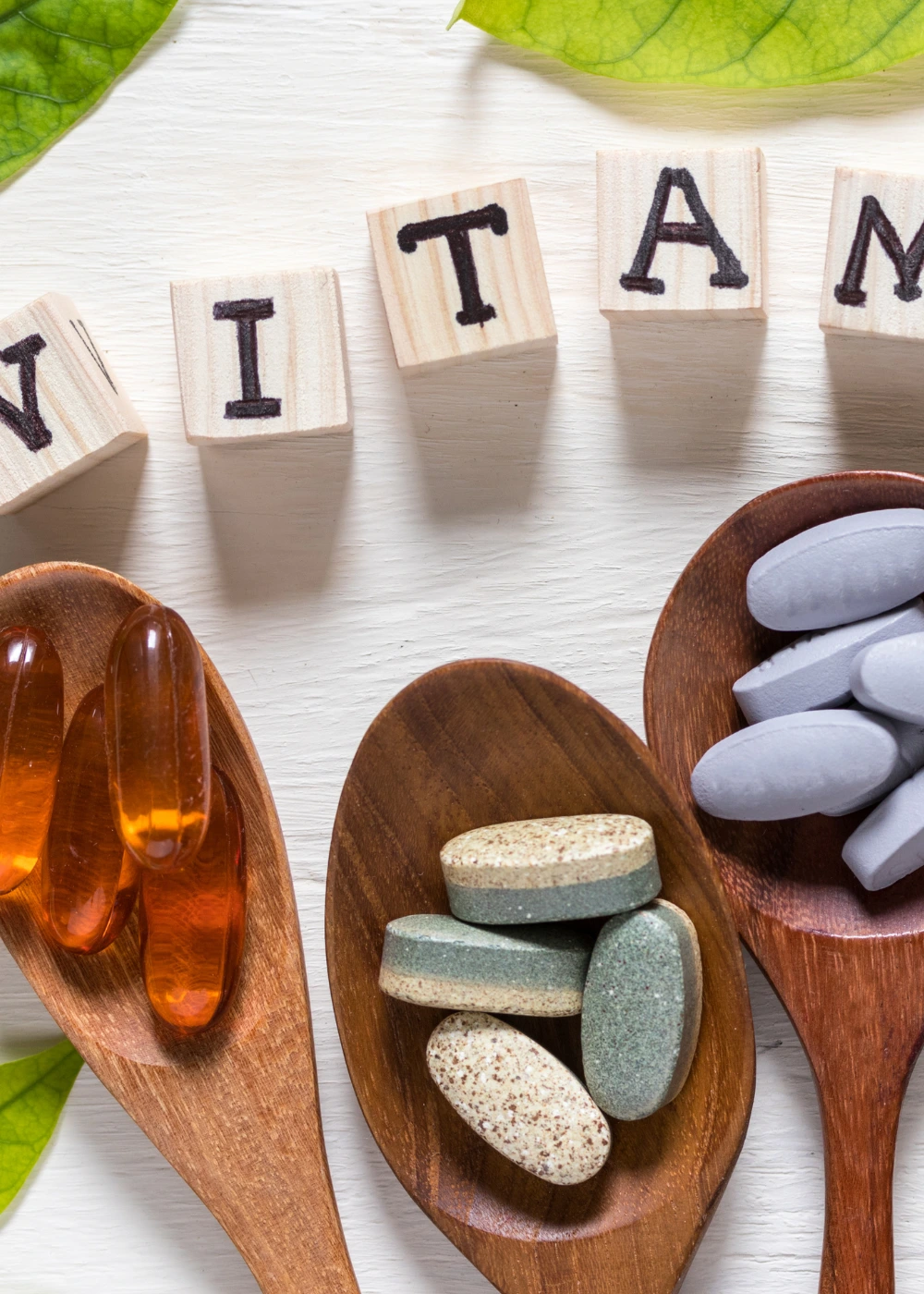 Wooden blocks spelling out "VITAMIN" are arranged on a light wooden surface, accompanied by three wooden spoons. One spoon holds orange gel capsules, another contains multicolored tablets, and the third has gray tablets, with green leaves placed on either side for decoration.