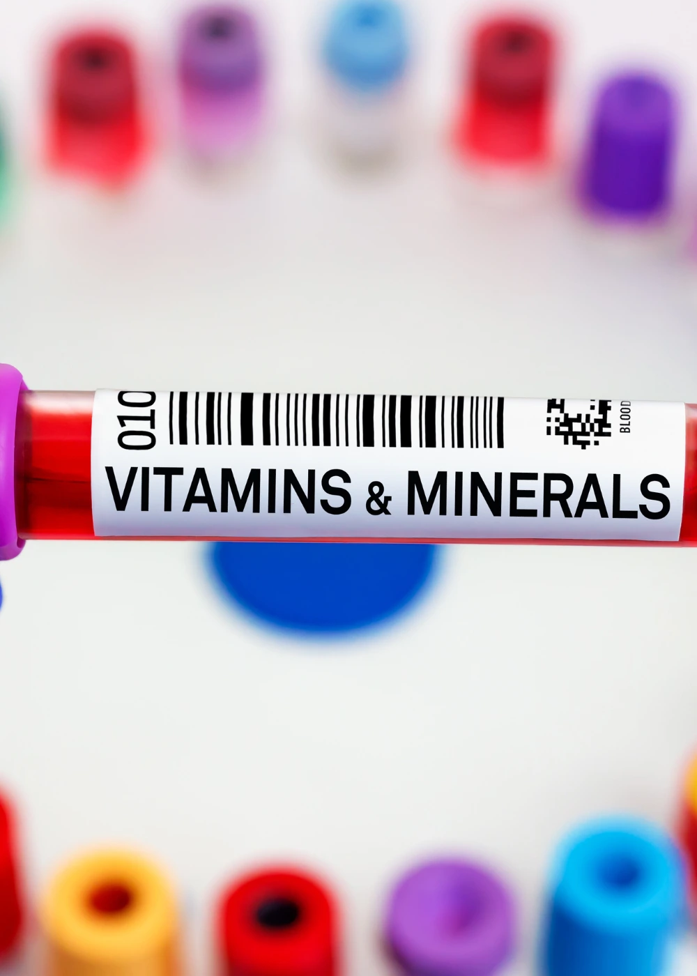 A test tube with a purple cap, containing red liquid and labeled "VITAMINS & MINERALS" with a barcode, is prominently displayed against a blurred background of other colorful test tubes in red, yellow, blue, and purple.