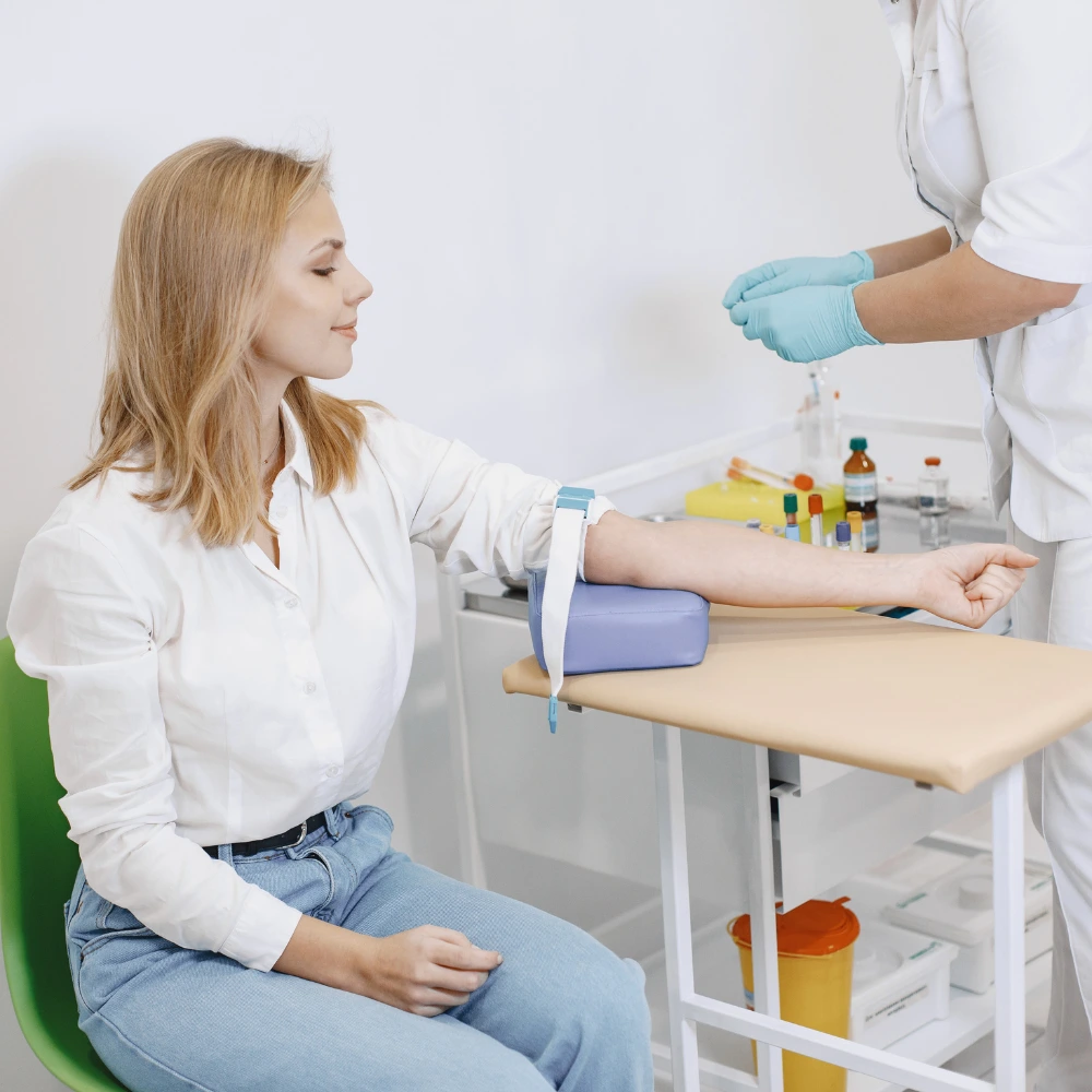 A woman in a medical setting, with her arm resting on a cushioned support while a healthcare professional prepares to draw blood.