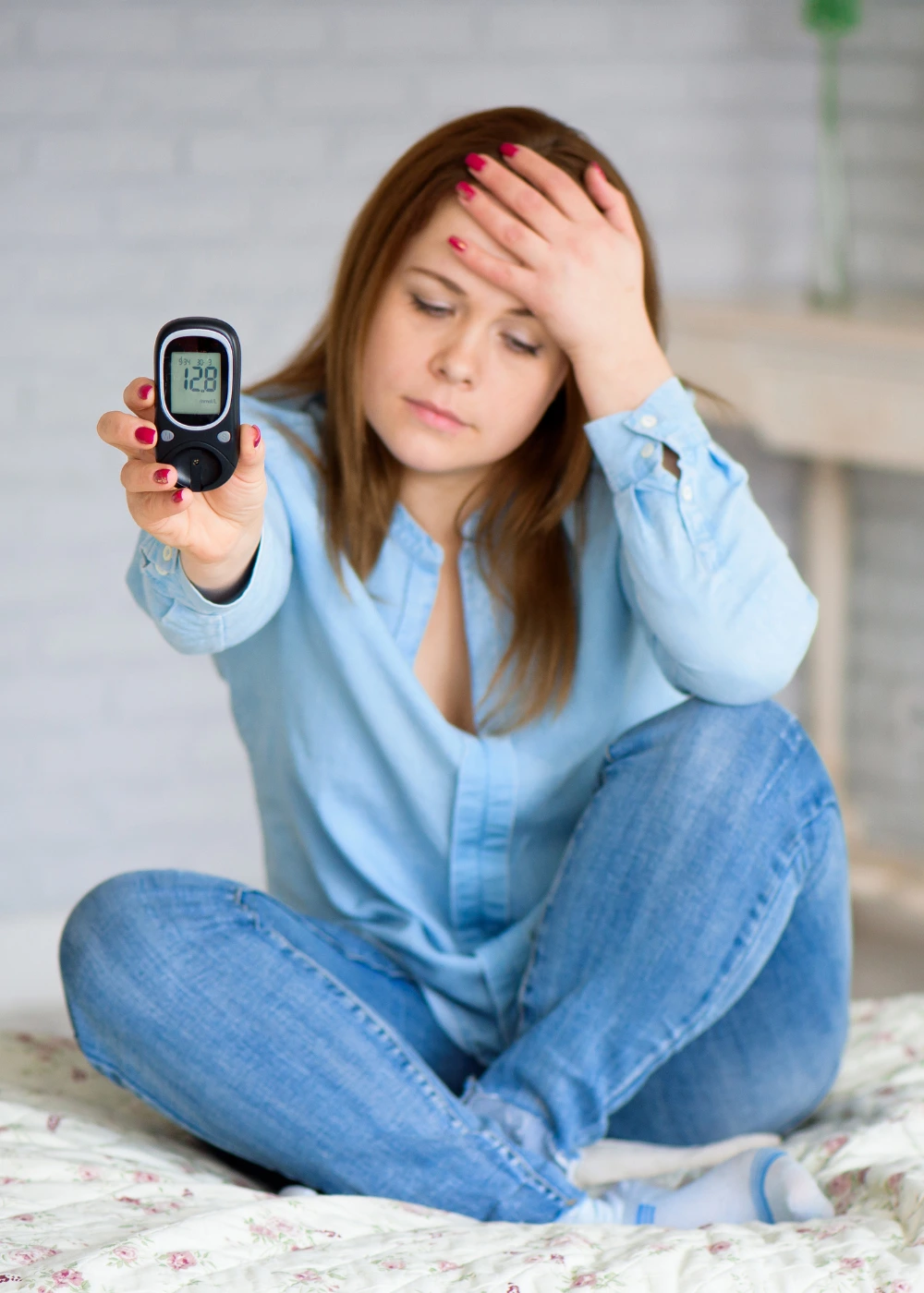 A woman sits on a bed, holding a glucose meter. She appears distressed, with her hand on her forehead.