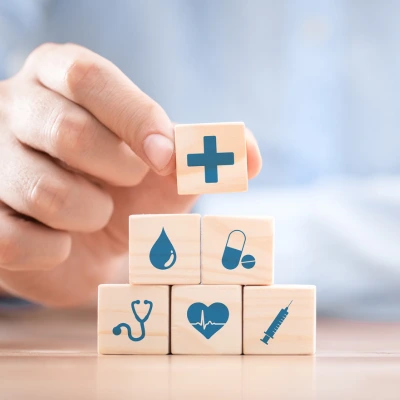 A hand is holding a wooden block with a plus sign on top of a stack of wooden blocks, each featuring healthcare-related