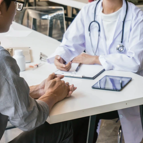 A patient consulting with a doctor at a clinic table, with a stethoscope, clipboard, and tablet visible, representing an urgent care medical consultation in Dubai.